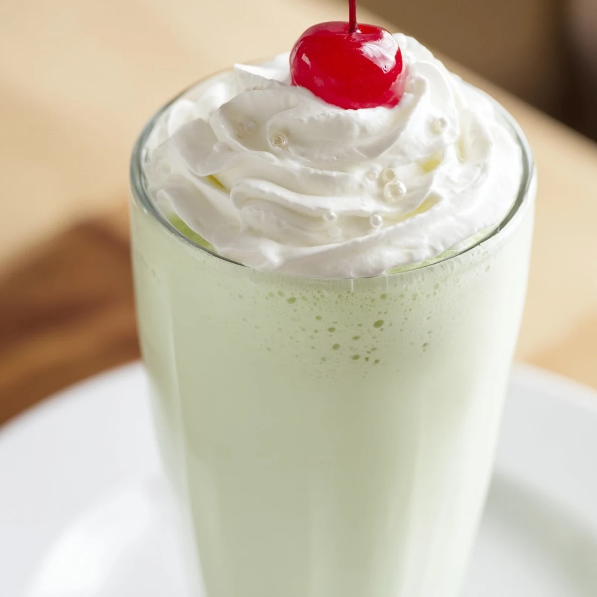 Two tall glasses of mint milkshake topped with whipped cream and maraschino cherries, garnished with fresh mint sprigs against a bright background.