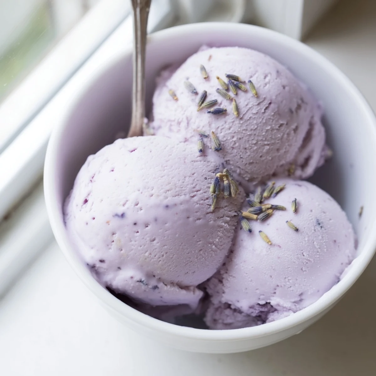 Scoops of Lavender Ice Cream sit in a chilled bowl, topped with fresh edible flowers and a sprig of lavender.