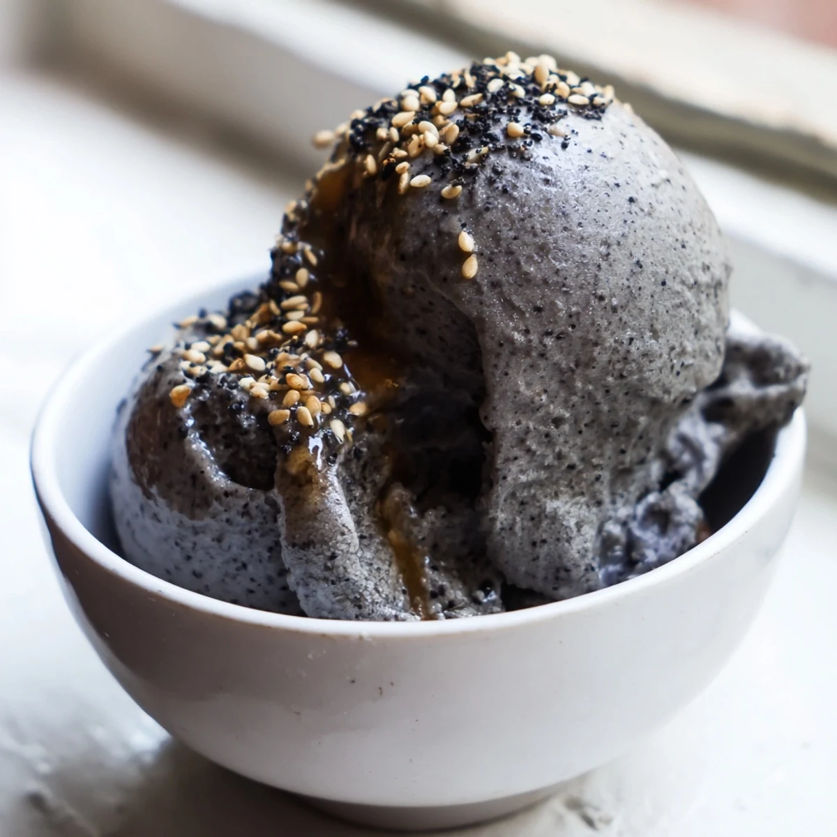 A rich scoop of Toasted Black Sesame Ice Cream in a chilled bowl, garnished with a drizzle of honey and toasted sesame seeds.  