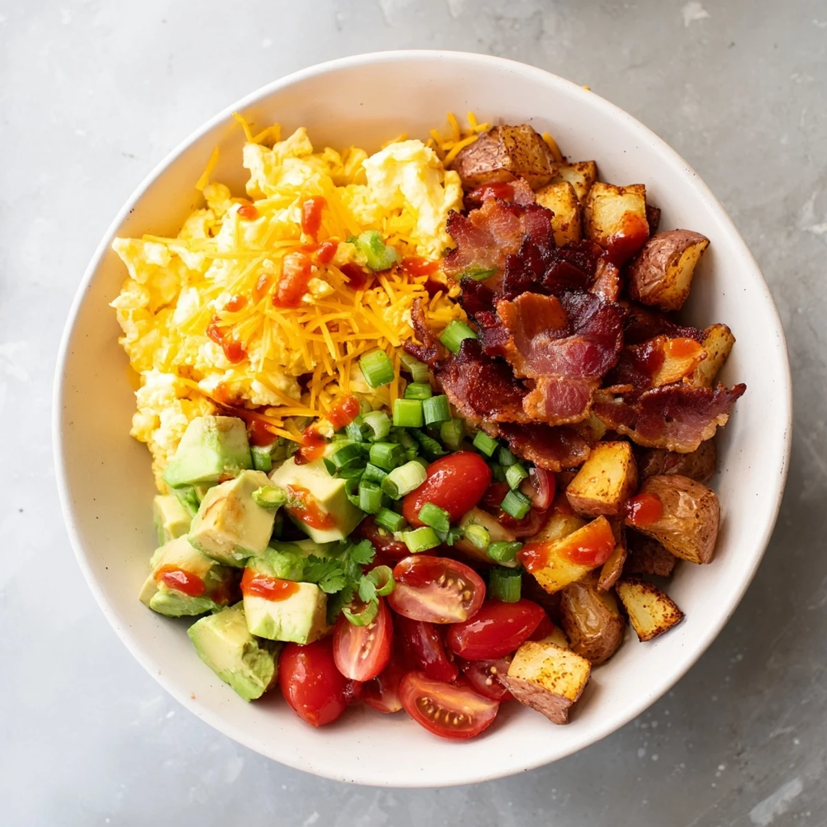 A close-up of a Loaded Breakfast Bowl showing golden crispy potatoes topped with fluffy scrambled eggs, crumbled bacon, and melted cheddar cheese.
